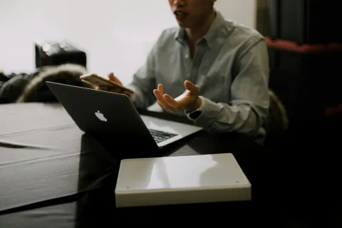 Person using a MacBook laptop and holding a smartphone while discussing at a dark table with a closed white box.