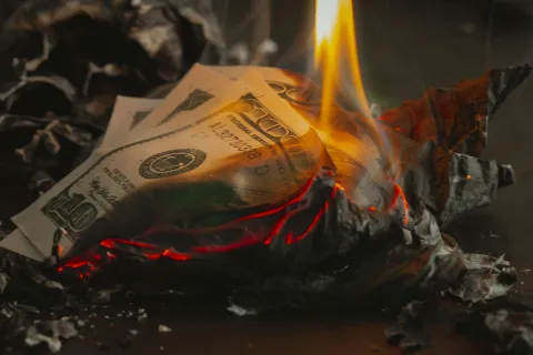 Burning US dollar bills with vivid flames and charred edges on a dark surface depicting financial loss.