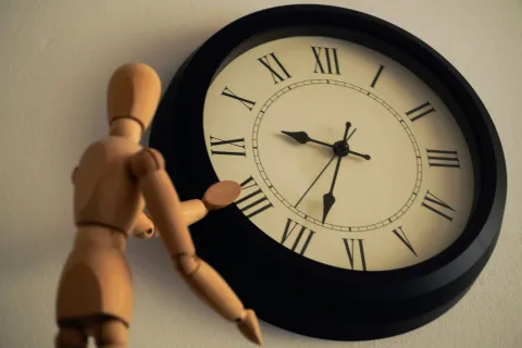 Wooden mannequin reaching toward large wall clock showing 5:45 with Roman numerals on cream face.