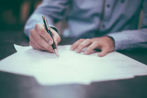 Person writing with a pen on multiple sheets of paper on a desk, wearing a light blue shirt.
