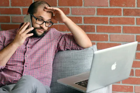 Man in glasses and checkered shirt looking stressed while talking on phone and using laptop on couch