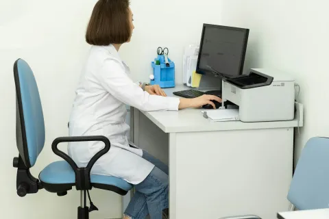Female doctor in white coat working at computer in a clean, modern medical office with blue chairs.