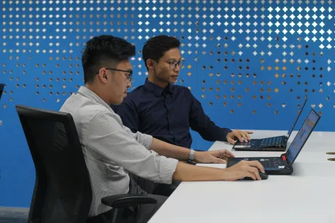 Two men working on laptops at a white table against a blue patterned wall in an office setting.