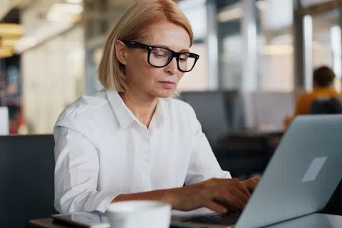 Mature woman wearing glasses focused on laptop in modern café with blurred background and coffee cup.