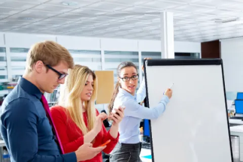 Three young professionals collaborate in office with blank flip chart and smartphones in hand.