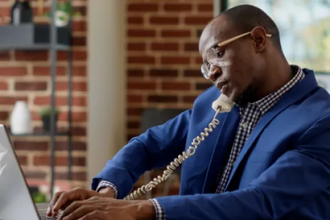 Man in blue suit using laptop and talking on corded phone in a modern office with brick wall background
