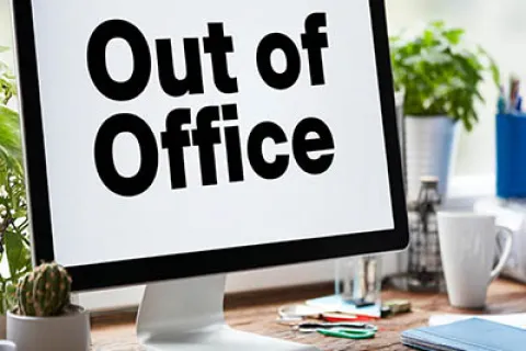Desktop monitor displaying Out of Office message on a wooden desk surrounded by office supplies and plants.
