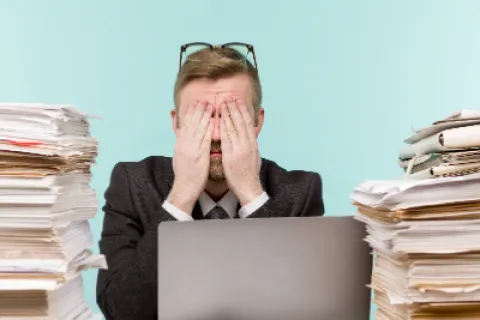 Stressed man in suit sitting at desk with laptop overwhelmed by tall stacks of paperwork on both sides.