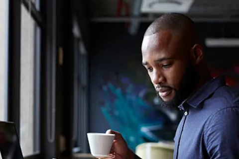 Man in blue shirt holding a cup and using smartphone at a wooden desk with an open laptop by a window.