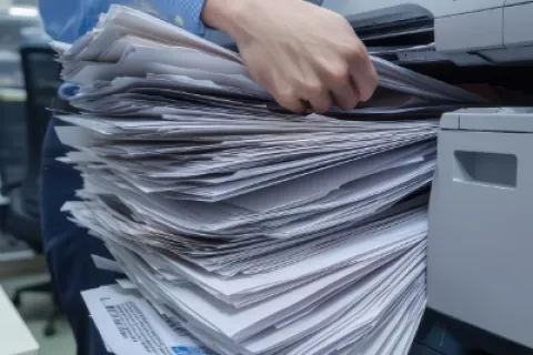 Person handling a large stack of printed papers coming out of an office printer in a busy workspace
