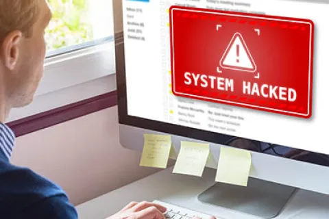Man at desk sees red system hacked warning on computer screen while typing on keyboard in office.