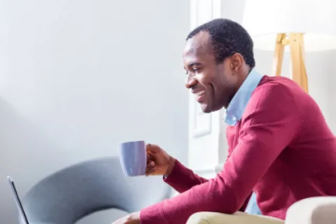 Smiling man in red sweater using laptop while holding a mug, seated in a bright modern room.