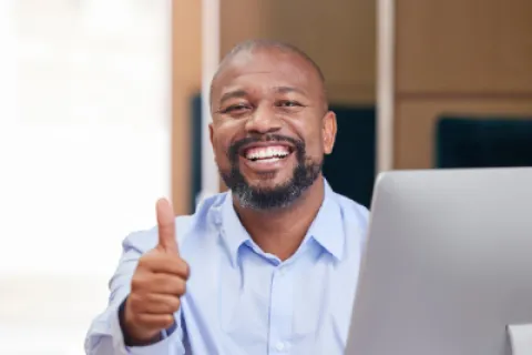 Smiling man in light blue shirt giving thumbs up while sitting at desk with computer and smartphone.
