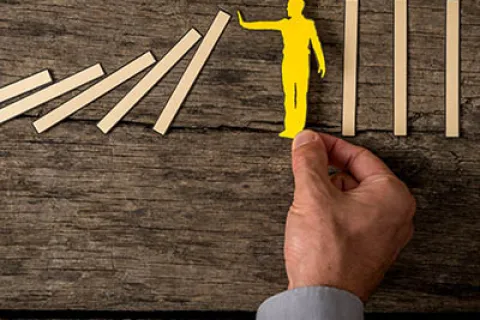 Hand holding a yellow figure stopping falling dominoes on a wooden surface symbolizing prevention and control.