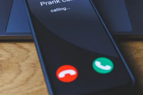 Smartphone screen shows incoming prank call with red decline and green accept buttons on wooden table.