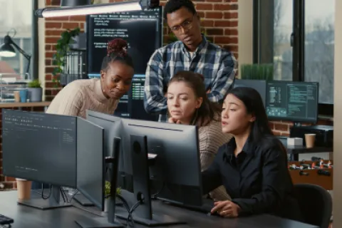 Four diverse coworkers collaborating and analyzing code on multiple computer screens in an office with brick walls.