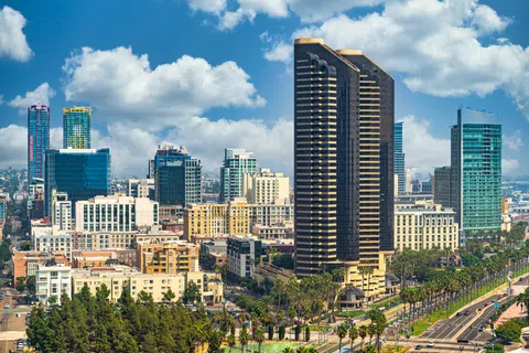 Cityscape of downtown San Diego with modern skyscrapers, palm trees, and a blue sky with clouds.
