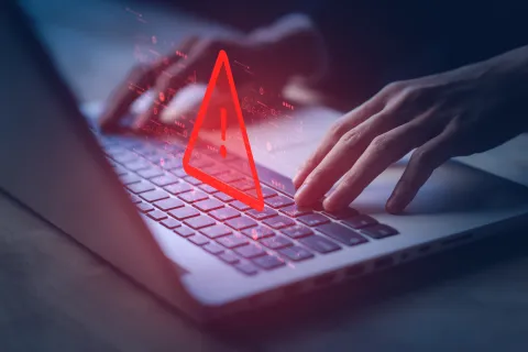 Hands typing on laptop keyboard with a red digital warning alert icon floating above it.