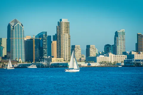 Sailboats float on blue water with a city skyline of tall buildings under a clear blue sky in the background.
