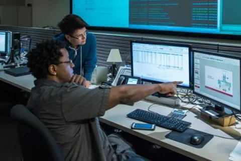 Two professionals analyzing data and schematics on multiple computer monitors in an office setting.