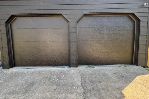 Two dark brown closed garage doors with angular trim on a house with horizontal siding and concrete driveway.