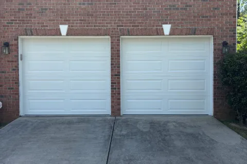 Two white garage doors on a red brick house with concrete driveway and a window above.