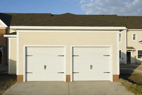 Two white garage doors with decorative handles on a beige house exterior under a clear blue sky.