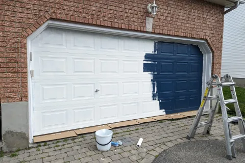 Half-painted white garage door with dark blue paint, ladder, paint bucket, and roller on driveway.