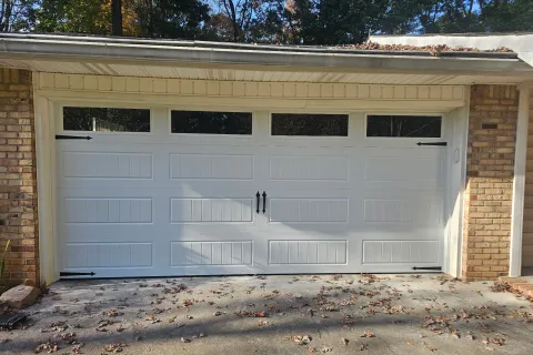 White paneled garage door with black hardware and windows at the top, set in a brick house exterior.