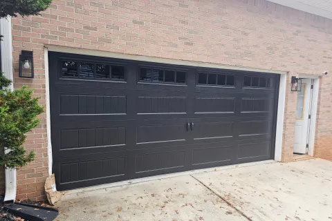 Modern black garage door with decorative windows installed on brick house with concrete driveway.