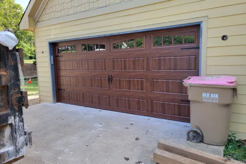 Wood-grain textured double garage door with windows and decorative iron handles on a yellow house.