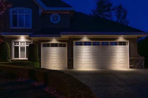 Suburban house with two illuminated garage doors at night and well-manicured bushes in driveway area.