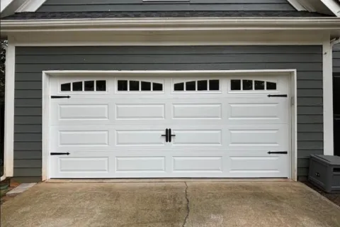 White garage door with decorative black hardware on a gray house exterior and concrete driveway.