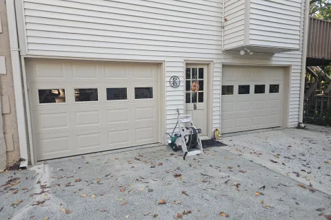 White siding house exterior with two beige garage doors, a central glass door, and scattered leaves on concrete driveway.