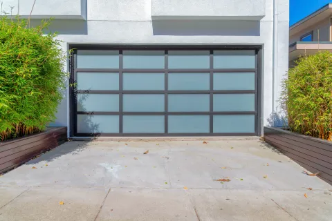Modern frosted glass garage door with black frame on a white house exterior and flanked by green shrubs.