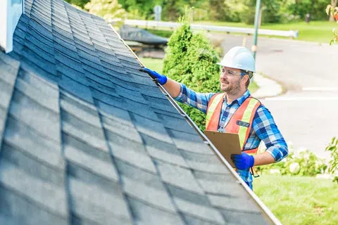 Construction worker inspecting roof shingles wearing safety gear and holding a clipboard outdoors.