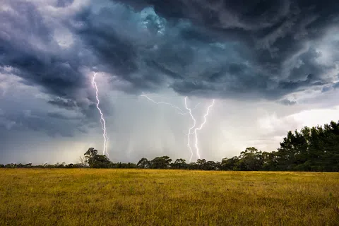 Lightning strikes over a wide grassy field under dark stormy clouds at dusk.