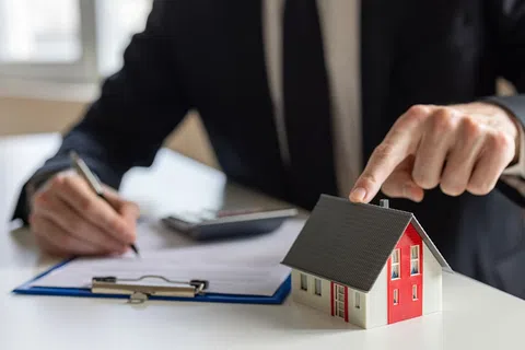 Businessperson pointing to a small house model on desk while signing a contract with a pen indoors.