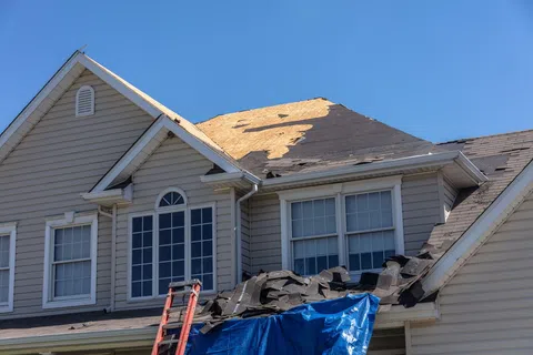 House undergoing roof replacement with shingles removed and a ladder leaning against the structure under clear blue sky.