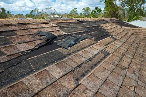 Roof with damaged and missing shingles showing exposed underlayment under a partly cloudy sky.
