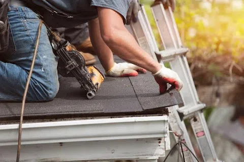 Worker installing asphalt shingles on a roof using a nail gun near a ladder in sunlight.