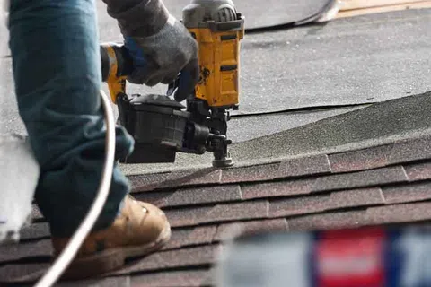Construction worker using a nail gun to install asphalt roof shingles on a house roof.