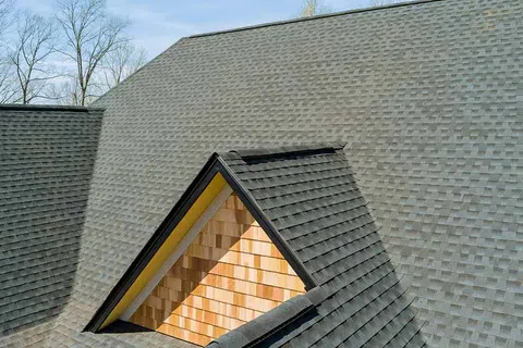 Close-up of gray shingled roof with a dormer window featuring wooden siding under clear sky