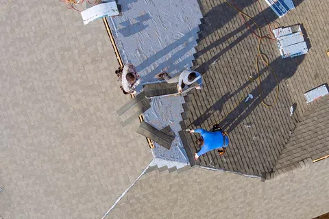 Aerial view of three workers installing roofing shingles on a house roof under sunny conditions.