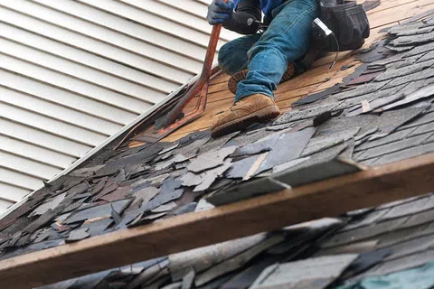 Roofer removing damaged shingles from a residential roof using a pry bar while wearing safety gear.