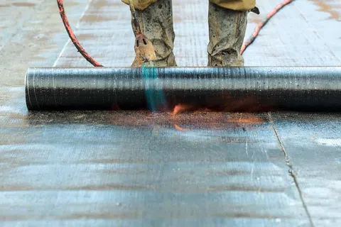 Worker using a blowtorch to apply waterproofing membrane on a flat roof surface wearing protective boots.