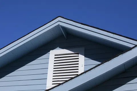 Blue house exterior with gable roof and white vent against clear bright blue sky