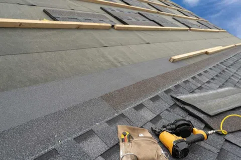 Roof under construction with asphalt shingles, wooden battens, nail gun, and tool pouch on a clear day.
