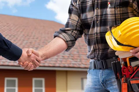 Construction worker holding a yellow hard hat shaking hands with another person in front of a house.