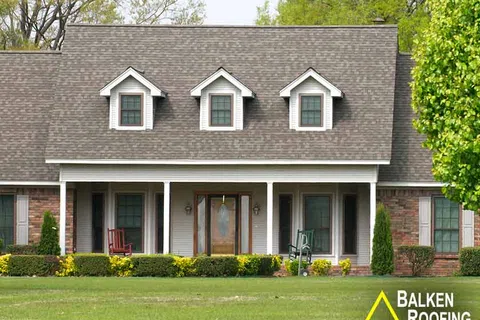 Traditional suburban house with gray shingle roof, three dormer windows, front porch, and well-kept lawn.